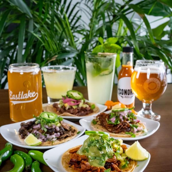 Four Sonora Grill tacos with various toppings, drinks, and a hot sauce bottle on a wooden table.