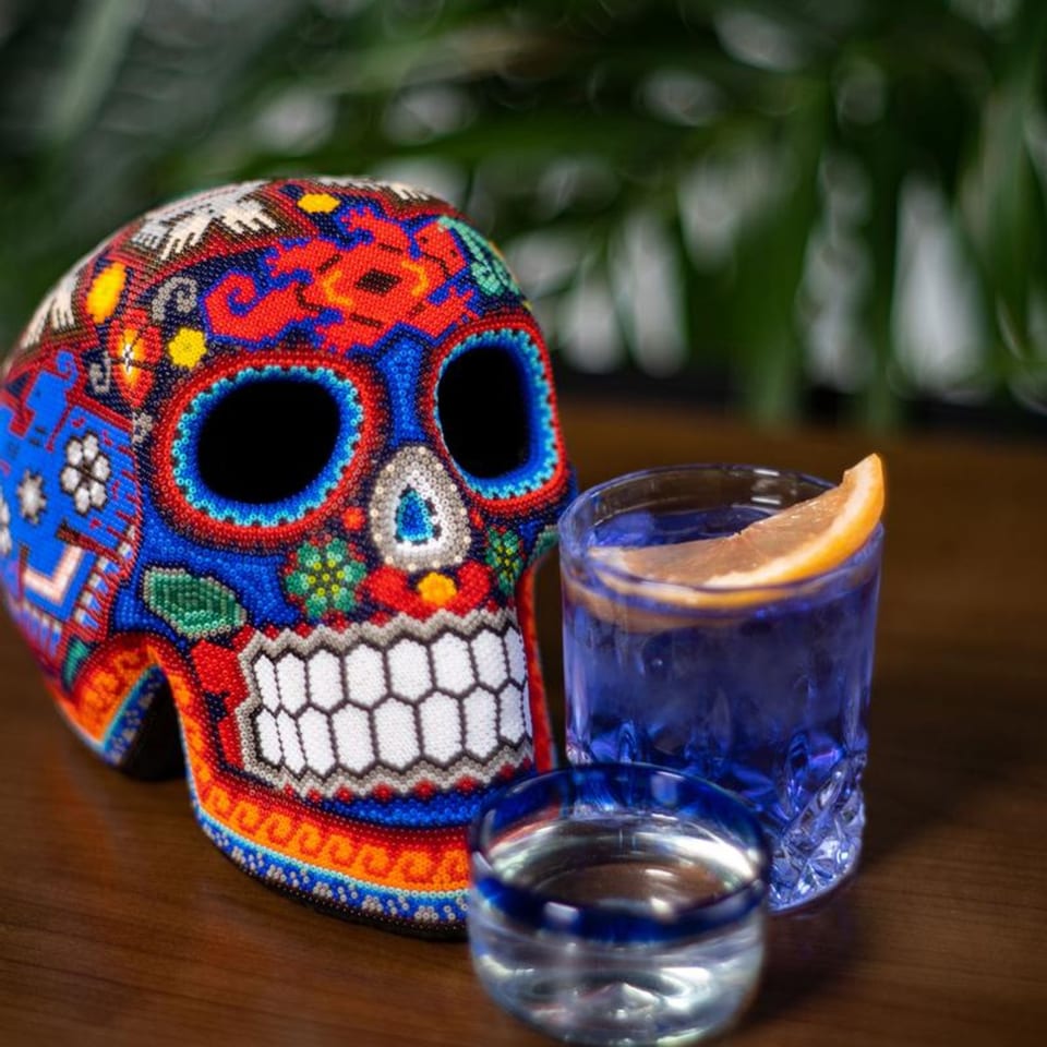 A colorful beaded Mexican sugar skull next to two glasses with blue drinks and orange slices.