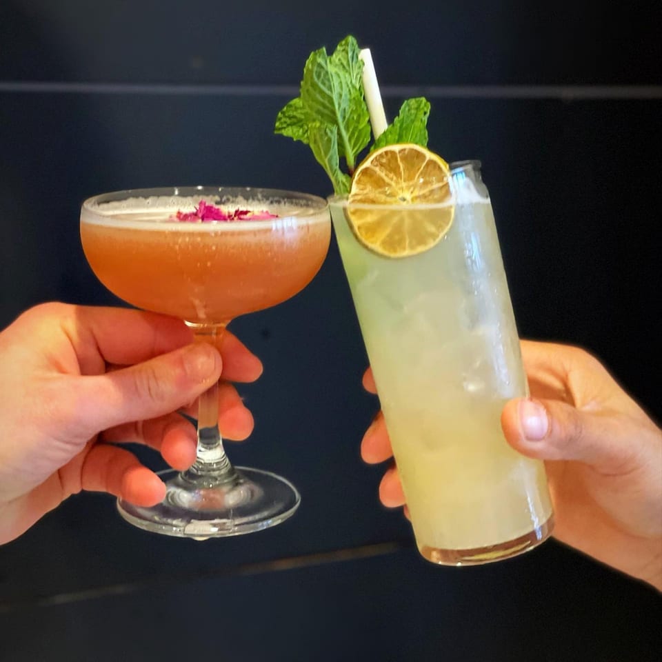 Two hands holding up cocktails, one with rose petals, the other with mint and lime.