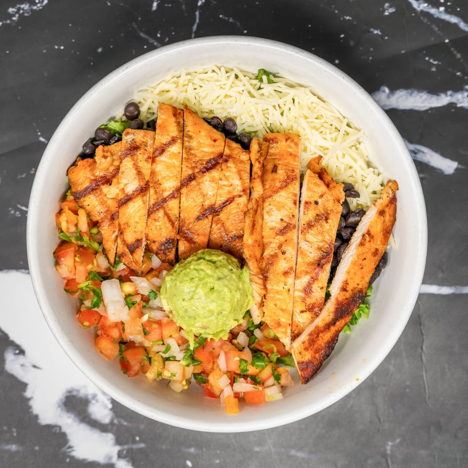 Grilled chicken, black beans, shredded cheese, pico de gallo, and guacamole in a white bowl.