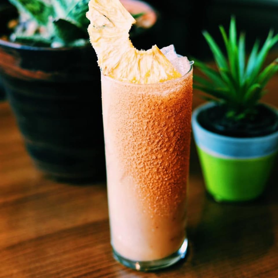 A tall glass of a light orange drink, rimmed with spices and garnished with dried pineapple.